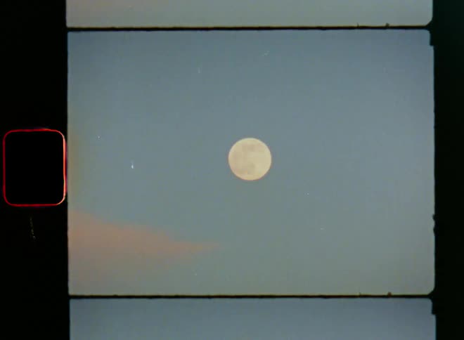 The full moon shining in a bright blue sky during evening time as the setting sun makes the clouds glow pink or red in the sky on vintage Super-8 film