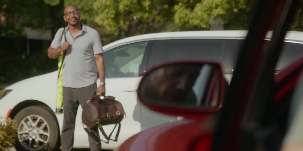 Man getting out of car with sports equipment greets a man bending into the front seat of a red car