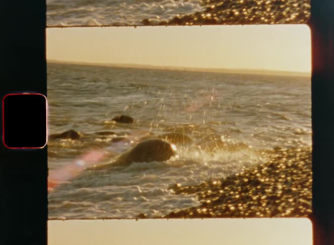 Ocean waves wash into a rocky coastline or gravel and splash around during the evening time of a sunny summer day caught on vintage film with bright lens flare and dynamic camera movement