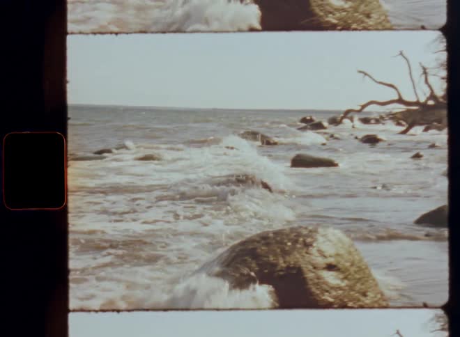 Waves crash against the shore and splash as they hit big boulders or rocks laying at the beach with vintage coastal vibes and ocean or sea flair