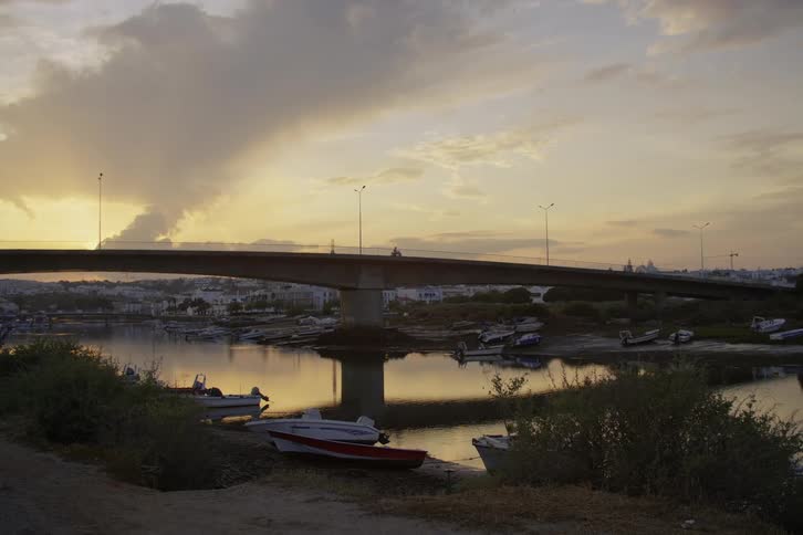 Motorcyclist crosses bridge over river lined by boats as sun shines from horizon