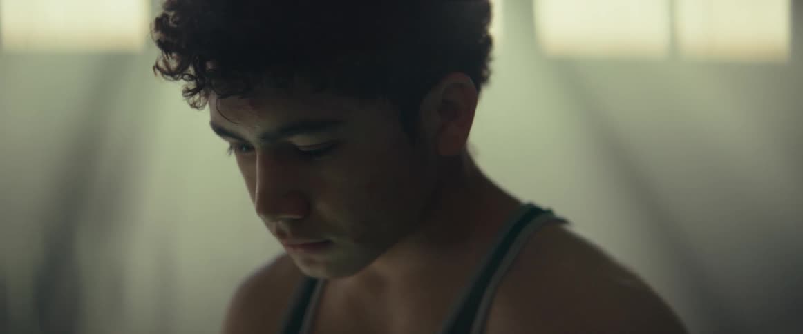 Youth wearing wrestling jersey stands in dimgym lit by rays from high windows