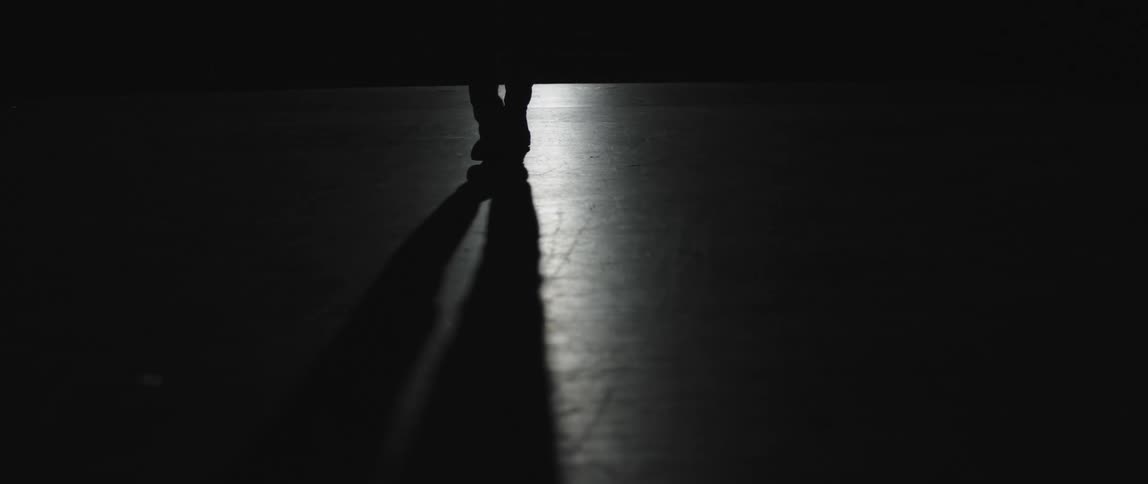 Feet of a person slowly walking through a dark room casting their body shadow ahead