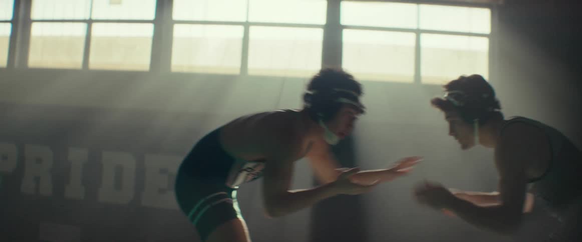 A pair of young men in wrestling singlets wrestle in a dimly lit gym during the day