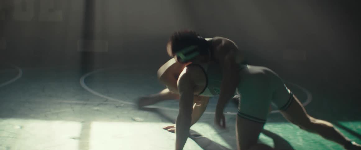 A pair of young men wrestle on a wrestling mat in a dimly lit gym during the day