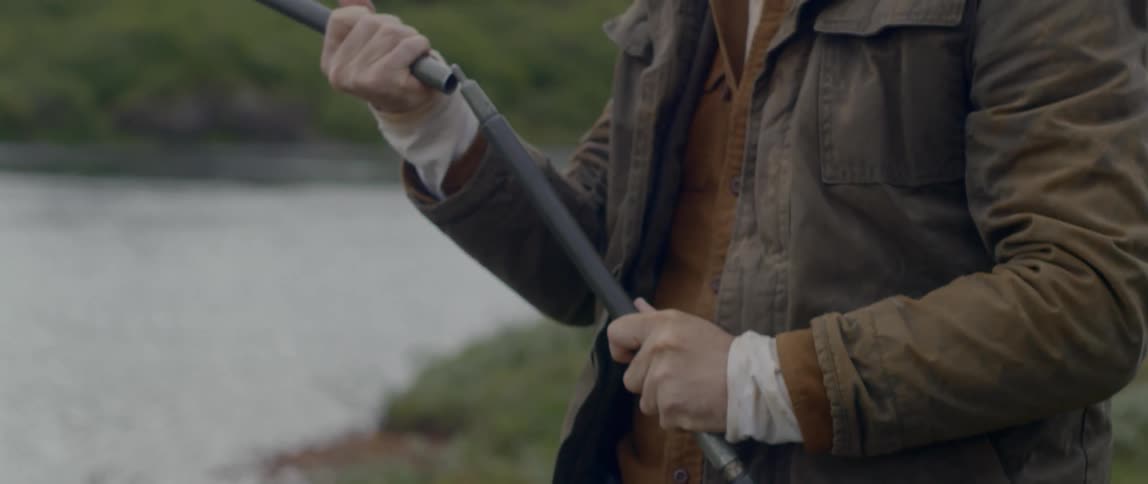 A man wearing a medium weight jacket is outdoors and next to a river as he leans over to pickup two metal rods and assembles them together