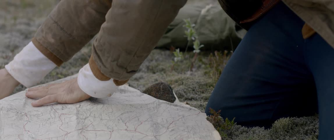A man uses stones to flatten out a map spread out on the grass