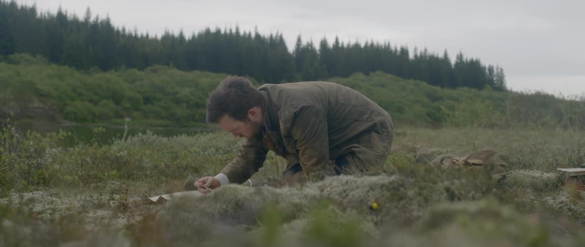 A well dressed man write something on a book in the middle of a grassland