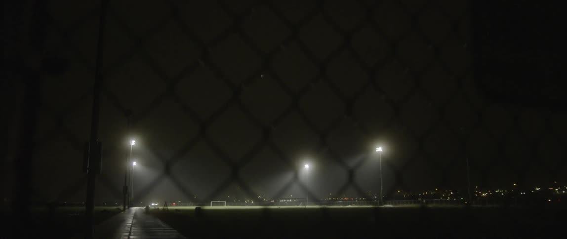 Behind the fence is a large field with lights in the distance