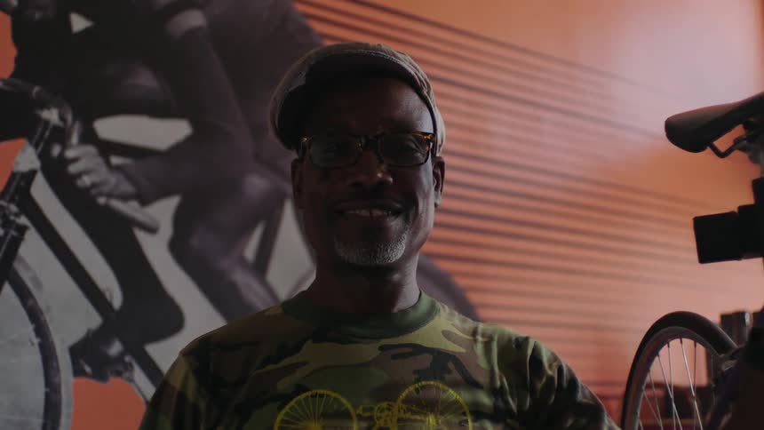 A man smiles into the camera while standing next to a bicycle in a bike shop during the day