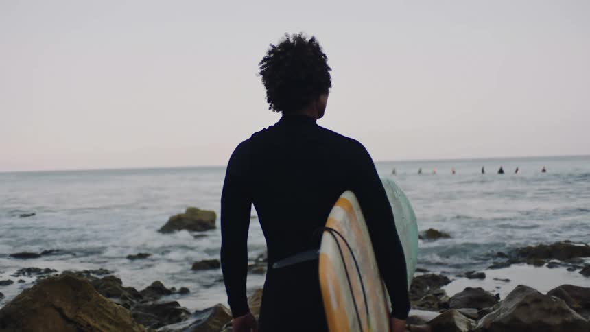 A surfer in a wet suit stands on a rocky coastline while carrying a surfboard