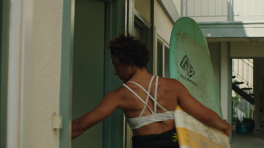 A woman carrying a surfboard exits an apartment and walks out of frame