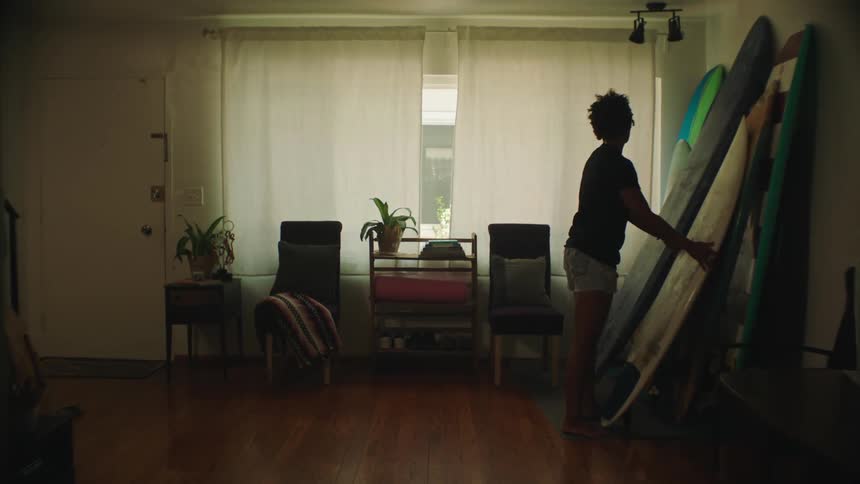 Woman walks across living room to a stack of surfboards against the wall and chooses one