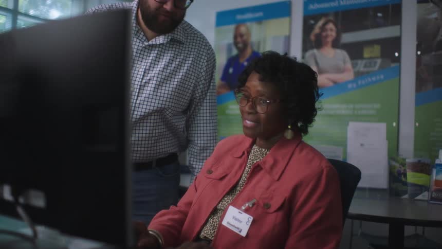 A man walks over to a woman sitting at a computer desk asking her something to which she smiles