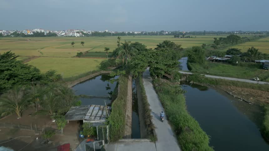 An aerial shot of a pedestrian road along a waterway between grassy fields and farms