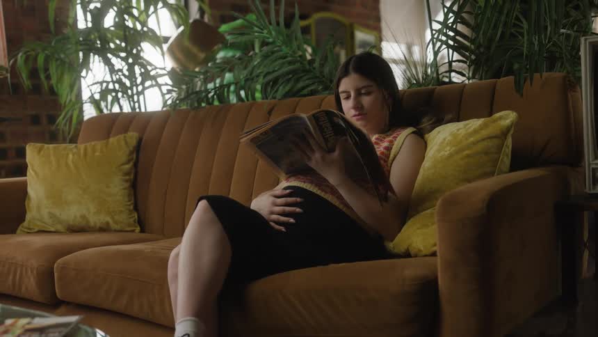 Pregnant woman sits on a couch backed by potted plants and reads a magazine