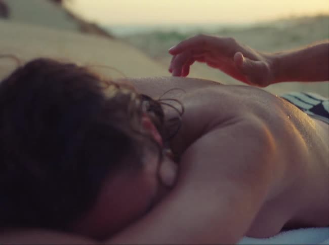A hand is touching a womans back as she lays facedown on the beach