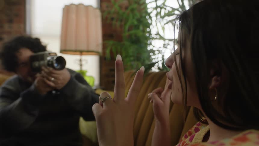 Man recording a woman posing happily for a photo with a camcorder both on the couch
