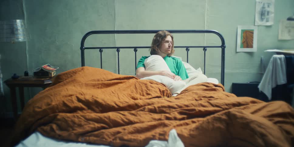A man is sitting up in bed frustrated as the camera zooms pushing him back to the wall