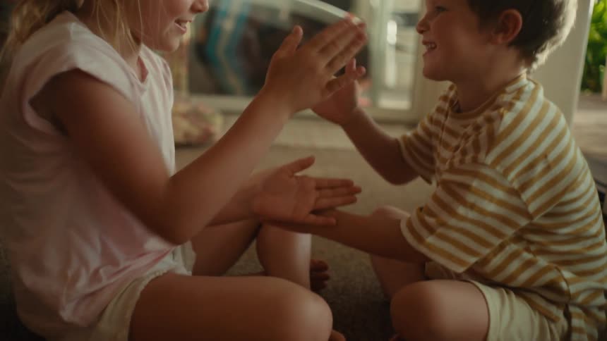 A girl and boy play a clapping game together while sitting on the floor indoors during the day
