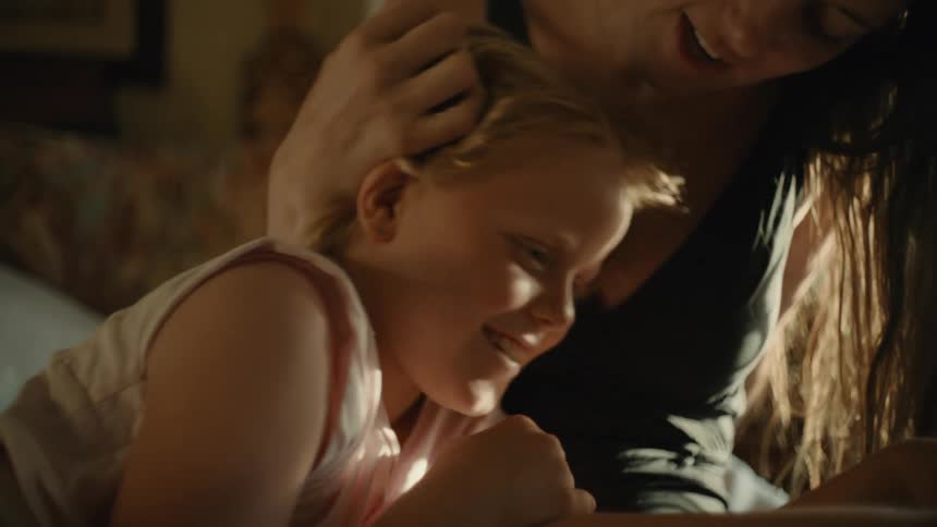 A woman and a young girl lying on a bed with the woman talking to the girl and caressing her head as the girl laughs