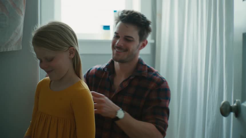 A man brushes the hair of a girl while kneeling behind her indoors during the day