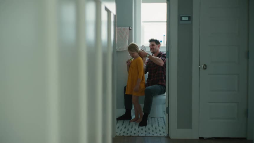 A man brushes the hair of a girl while she brushes her teeth in a home bathroom during the day