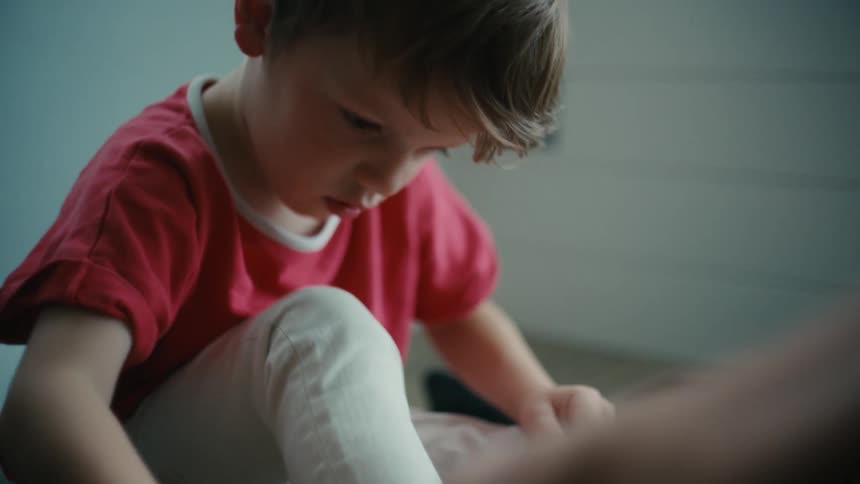 An adult helps a young boy to tie his shoes while they sit on the floor indoors during the day