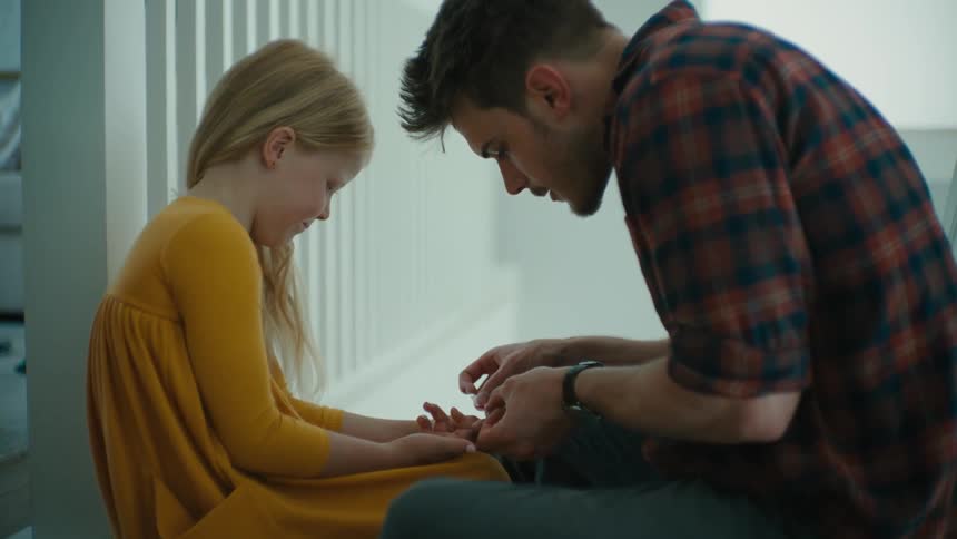 A man applies an adhesive bandage to the finger of a girl while they sit at the top of a staircase indoors during the day