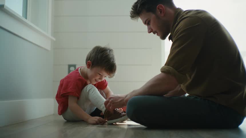 A man helps a boy to tie his shoes while they sit together on the floor indoors during the day