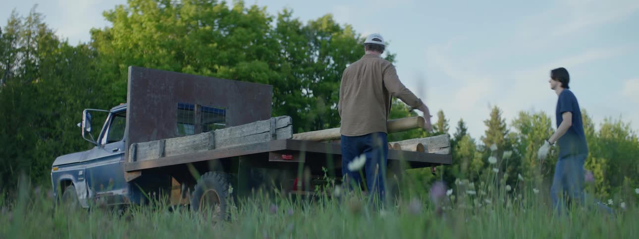 A pair of men retrieve logs from the back of a truck and carry them out of frame
