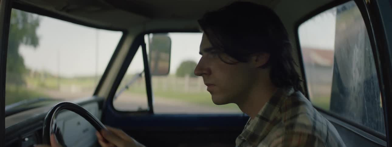A man is sitting in the driver seat of a truck turning the steering wheel as he drives around on a farm