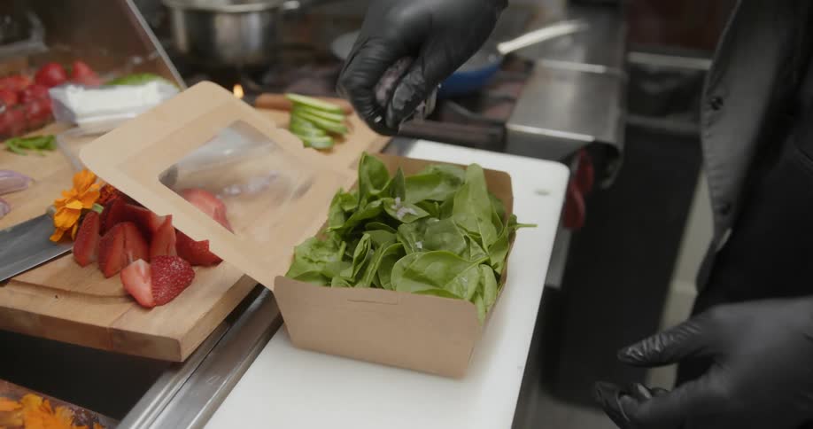 A close up of a person wearing black gloves pouring ingredients over a salad in a box