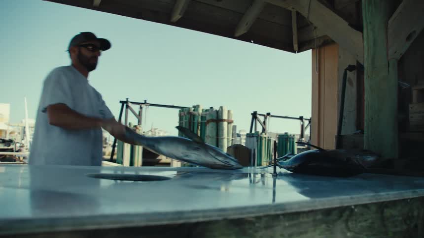 Man places fish on stone counter in porch near field crowded with tanks and people cooking outdoors