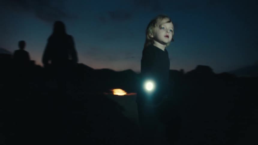 A little boy holding a flashlight running around in the dark near adults with a fire in the background