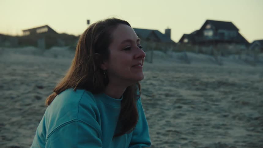 Woman sits on beach with houses in the background and wipes her face as she cries