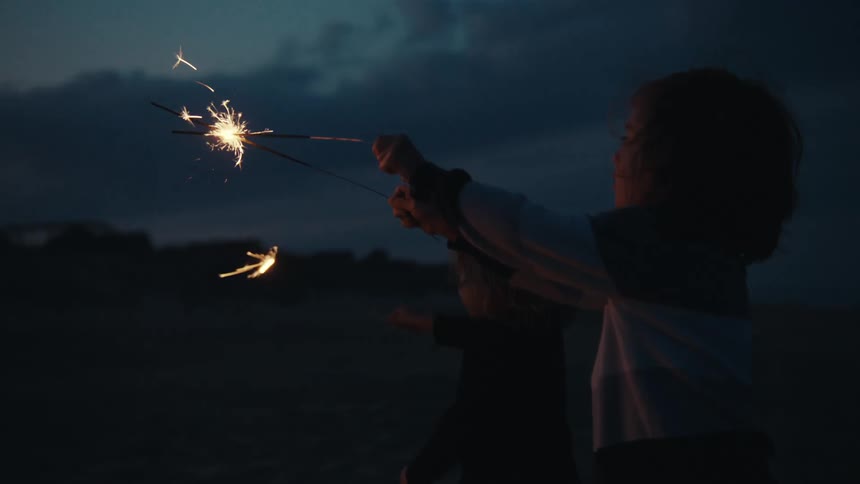 Two little boys holding sparklers in their hands as they stand near a fire on the beach