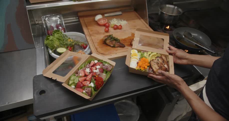 Female chef places salads in to go boxes on counter near stove
