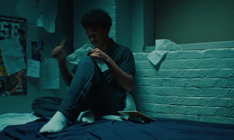 A young man in a jail cell rips off a page from a notebook then reads it
