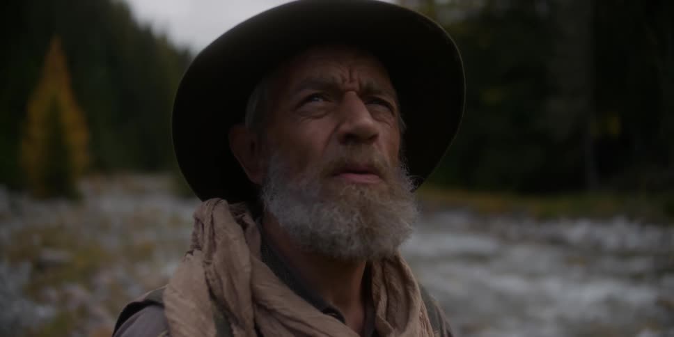 An older man standing up from a crouched position near a rivers edge as he looks up contemplatively