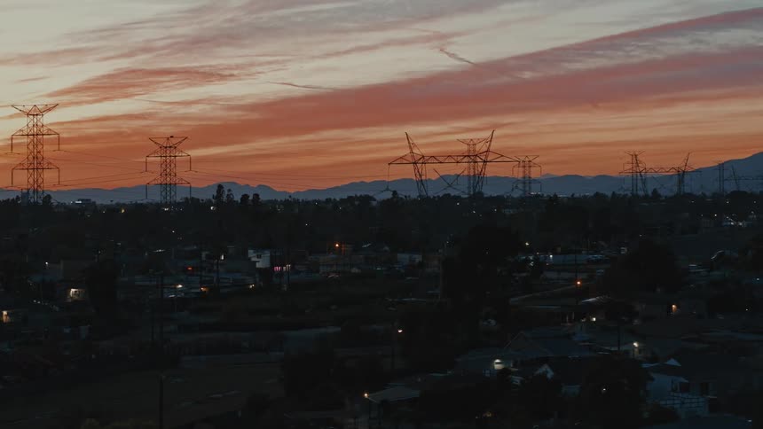 Electric transmission towers stand silhouetted against red sky as traffic passes through neighborhood nearby