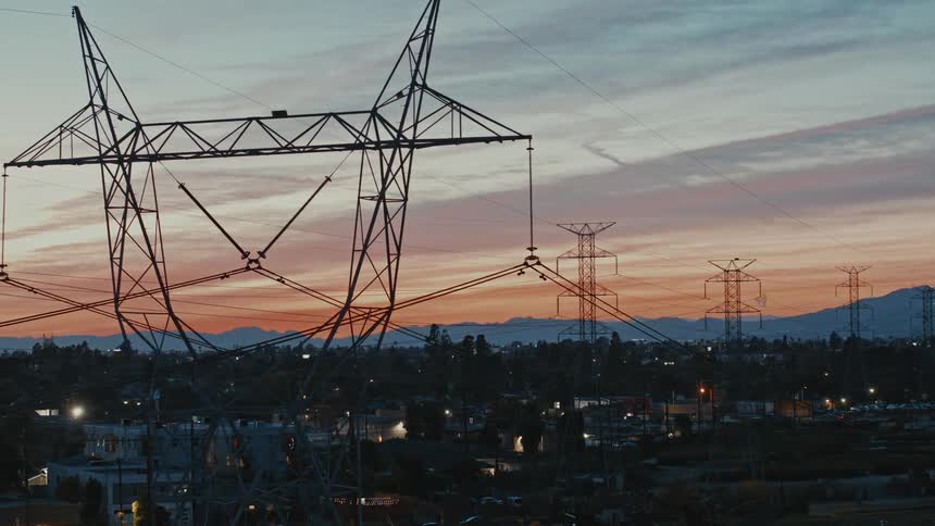A row of transmission towers loom over a residential neighborhood at sunset