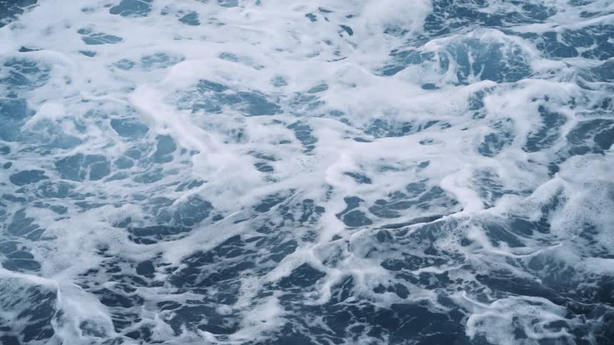 Drone view of ocean waves crashing up against rocks creating foam and ripples across the surface