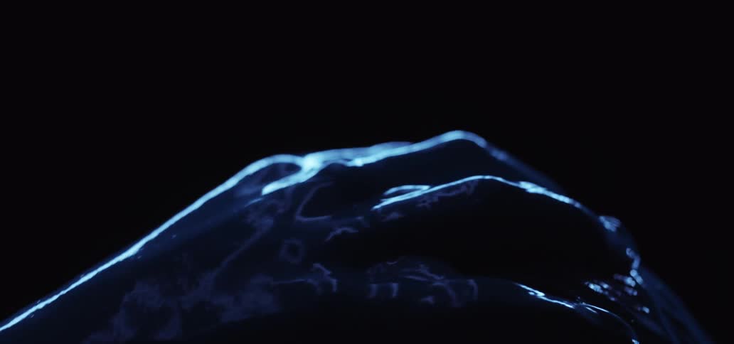 Person strokes blue paint covered hand over blue paint covered hair and forehead against pitch black background