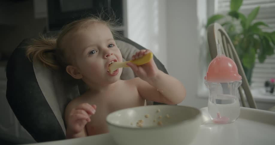 Blonde toddler sitting in high chair feeds herself with spoon