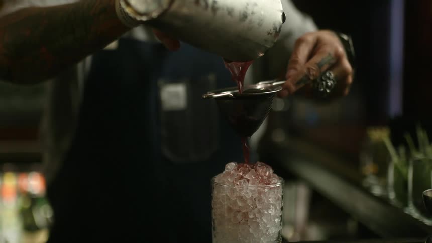 A bartender pours juice from a cocktail shaker through a strainer and into a glass filled with ice chips