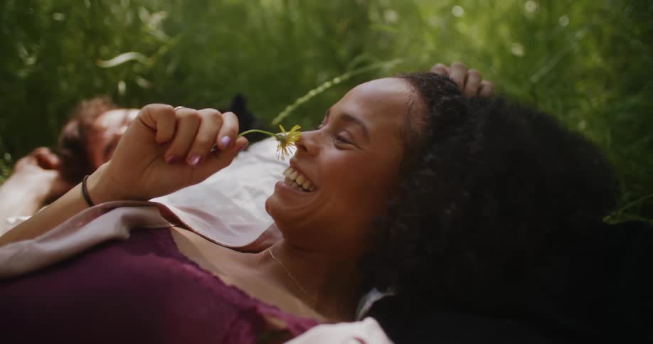Woman reclining against man as they both lie in field brings flower to her nose before smiling