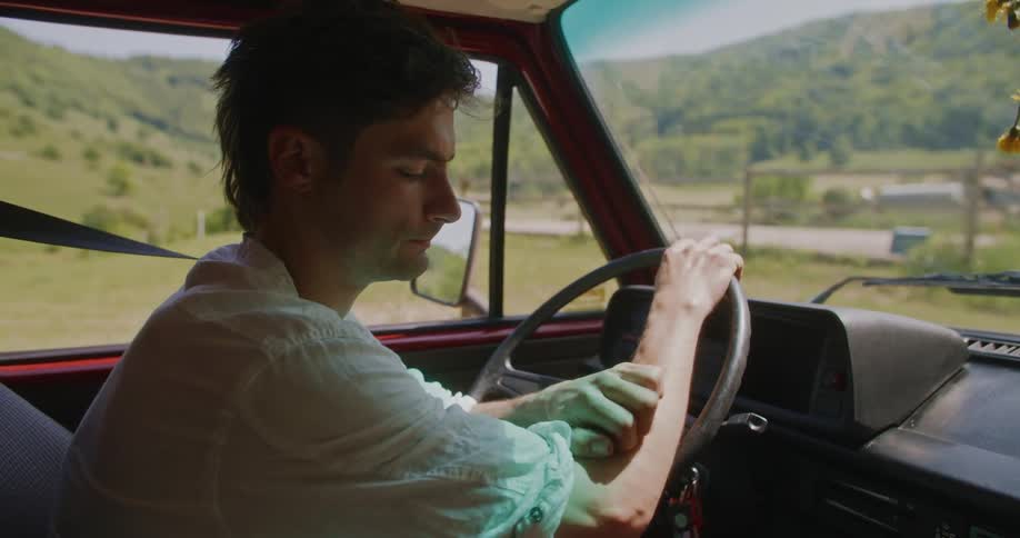 Man scratches his arm while sitting in drivers seat of van parked near road through mountains