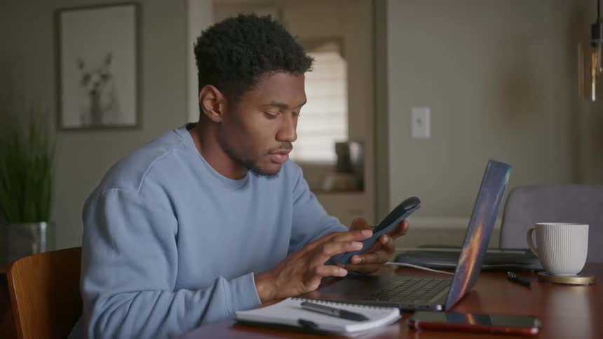 Man sitting at a table typing into a calculator in his hands before setting it on a laptop and furrowing his brow