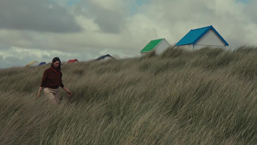 A woman walks through the tall grass in front of colourful beach houses